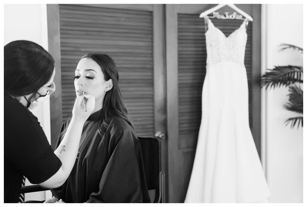 Bride getting makeup done with her white dress hanging behind her
